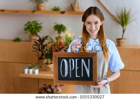 beautiful young florist in apron holding sign open and smiling at camera in flower shop