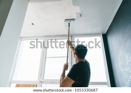 the man paints the walls and the ceiling in gray color, standing with his back to the camera. Focus on the roller. Painting and repair of the room. Royalty-Free Stock Photo #1055330687
