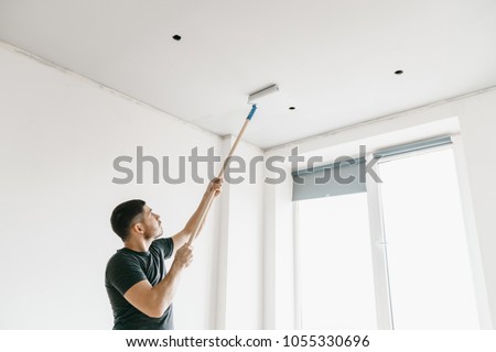 a man in a gray T-shirt with a roller on a long stick paints the ceiling in gray at home. Repair of the premises. Royalty-Free Stock Photo #1055330696