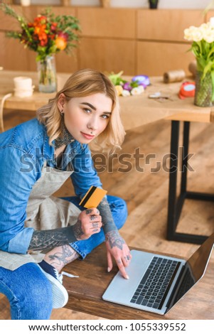 young florist holding credit card and looking at camera while using laptop in flower shop 