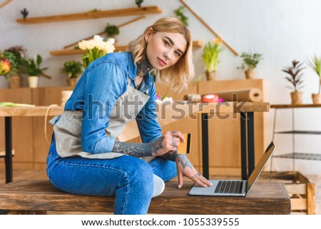 beautiful young florist holding credit card and looking at camera while using laptop in flower shop 
