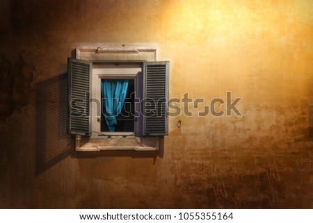 An open wooden old window on a plastered stone wall with a blue curtain, lit by light from a night street lamp.
