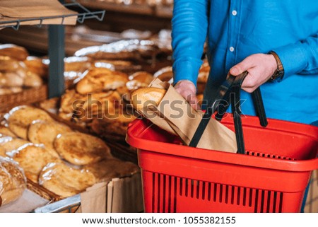cropped shot of man putting loaf of bread into shopping basket in supermarket