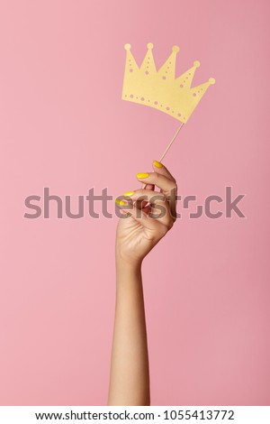 Female hand with yellow manicure hold a paper  craft crown on a stick. Isolated on a pink background. Beauty salon concept.