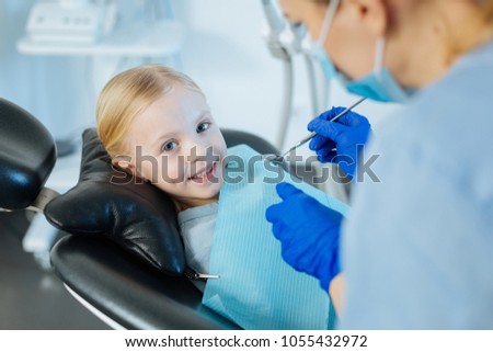 Nice treatment. Upbeat little girl lying in a dentist chair and smiling at the camera during a professional dental treatment carrying out by a female dentist