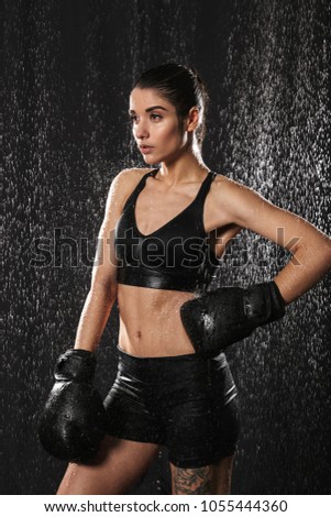 Photo of wet attractive woman with hair in ponytail putting arm in boxing gloves on waist and looking aside under water drops isolated over black background