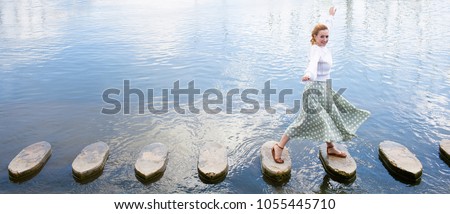 Panoramic view of joyful young woman playful stepping on stone steps crossing water in coastal destination on holiday, sunny outdoors. Fun female looking, travel recreation adventure leisure lifestyle Royalty-Free Stock Photo #1055445710