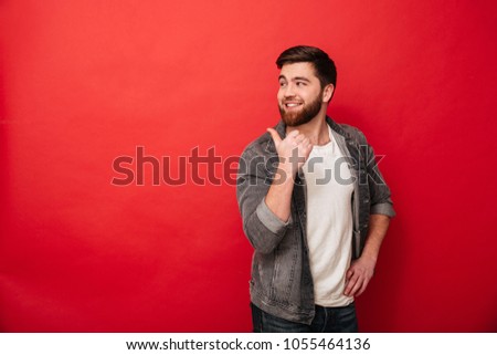 Photo of handsome man 30s in casual clothing gesturing fingers backward on copyspace with happy smile isolated over red background