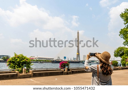 woman traveler with bag cross body and hat and take a photo with smartphoneat “Rama eight Bridge” background from Bangkok Thailand. Traveling Thailand.