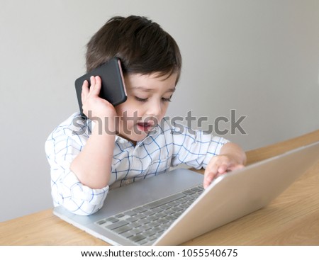 Busy little boy talking on the phone while typing on keyboad, Smart kid watching cartoon on laptop and speaking on mobile phone. Children development and Technology concept