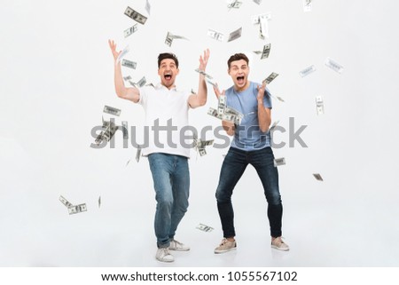 Full length portrait of two happy young men standing under money banknotes shower and celebrating success isolated over white background