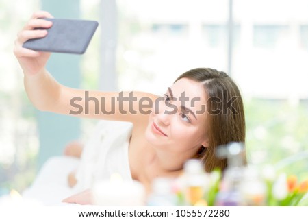Young Caucasian attractive woman lying on bed at spa salon, smiling and relaxation. Taking selfie photo from smartphone while waiting having body oil massage at spa resort with spa equipment.