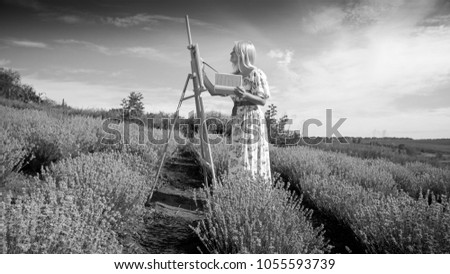 Black and white photo of young woman in long dress painting picture of field