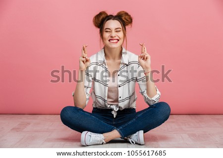 Portrait of a smiling young girl sitting on a floor with crossed legs and holding fingers for good luck isolated over pink background Royalty-Free Stock Photo #1055617685