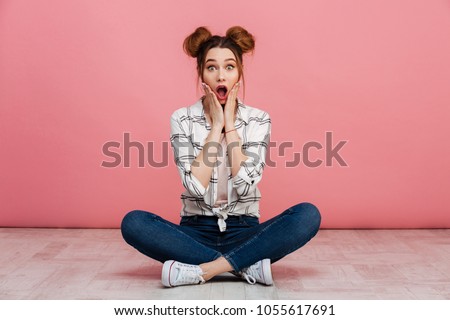 Portrait of a shocked young girl sitting on a floor with crossed legs isolated over pink background