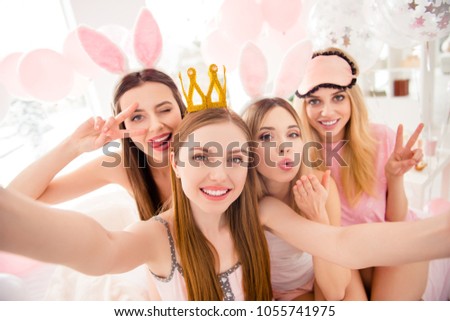 Attractive, charming, cheerful, stylish girls with crown, bunny ears, eye mask shooting self portrait on front camera showing two fingers, sending kiss with palm,  having theme party, dress code