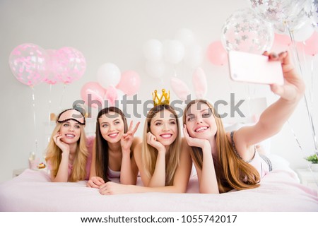 Cheerful, joyful, charming, pretty, funny, dreamy, cute models lying on bed shooting self portrait on front camera, holding using smart phone, showing two fingers, peace symbol, enjoying theme party