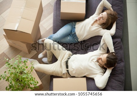 Relaxed smiling couple resting on couch moved in new home with boxes, happy real estate renters owners relaxing on sofa in living room, easy relocation with moving delivery service concept, top view Royalty-Free Stock Photo #1055745656