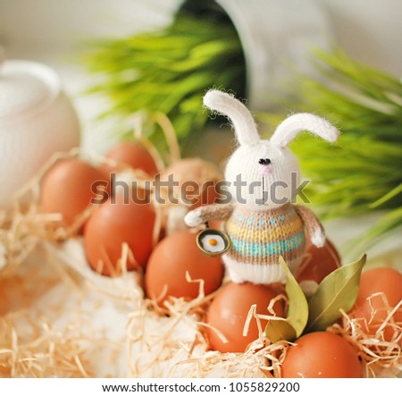 Handmade knitted toy. Easter bunny in a colored sweater holding a frying pan with eggs in his paw against the background of boxes with eggs