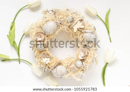 Easter wreath with straw, eggs and other traditional elements and white tulips around. Top view image of a pastel colored hand made home decoration for the spring holidays.