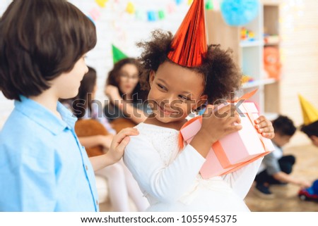 Nice little girl in white dress is pleased with gift she was given by boy in blue shirt at birthday. Happy birthday party. Gift time.