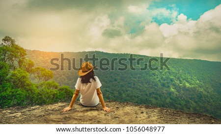 Young traveling woman wearing hat and sitting on the top of the mountain cliff with relaxing mood and watching beautiful view of woods and blue sky and clouds on vacation. Asian woman travel alone. Royalty-Free Stock Photo #1056048977