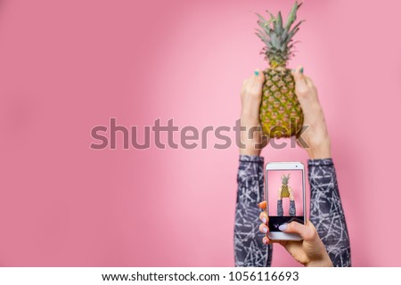 Sport, fitness, lifestyle, technology and people concept - young woman with smartphone taking selfie in gym while holding pineapple with her legs over pink background