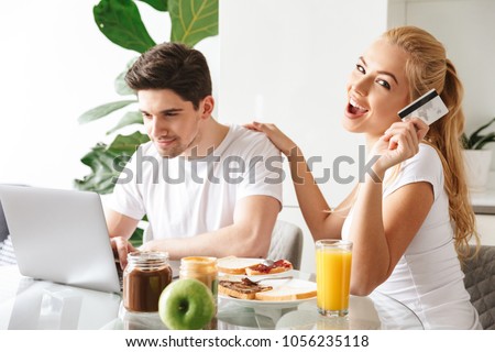 Photo of cute smiling young loving couple have a breakfast indoors at home while using laptop computer holding credit card.
