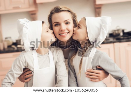 Children in chef's hats with mother in kitchen. Brother and sister are kissing mother on cheeks.