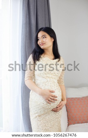 Beautiful young Asian pregnant woman standing by the window and touching her tummy. Pregnancy. Health. Motherhood. A cozy portrait of a pregnant woman.