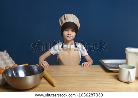 Picture of determined excited little boy wearing chef uniform standing at kitchen table with metal bowl, rolling pin, tray, flour and eggs, ready to start making dough for pancakes. Food and nutrition