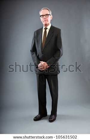 Expressive good looking senior man in dark suit against grey wall. Wearing glasses. Funny and characteristic. Well dressed. Studio shot.