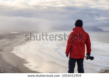 Adventurous man with a camera is standing taking pictures of the beautiful beach during a vibrant winter sunrise. Taken in Cape Kiwanda, Pacific City, Oregon Coast, America.