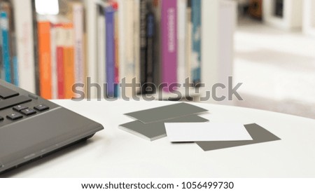 White and grey blank business cards and a laptop on a table in front of a blurred bookshelf in a sunny modern office ambiance, nobody