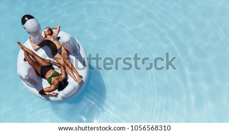 Overhead view of two women in bikini lying down on an inflatable toy in swimming pool. Girls sunbathing on floating pool inflatable toy.