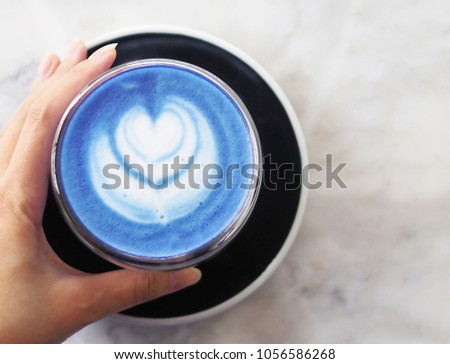 Top view of a woman's left hand grabbing a cup of hot butterfly pea latte with beautiful heart-shaped latte art on a light marble table background, morning refreshment