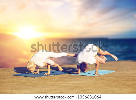fitness, people and healthy lifestyle concept - couple doing yoga side crane pose on mat outdoors on wooden pier over sea background