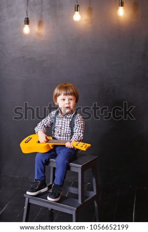 happy boy in a photo Studio with a black wall and lamps, in a plaid shirt with green suspenders and blue pants plays a yellow guitar and sings