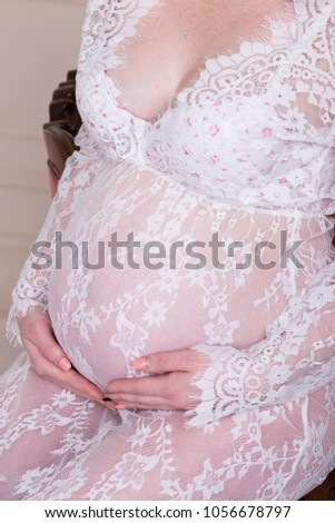 Close up of pregnant woman belly. Sitting and touching her belly in white lace dress. Pregnant woman body. Her hands on stomach, hands over tummy. Motherhood, pregnancy and expectation concept.