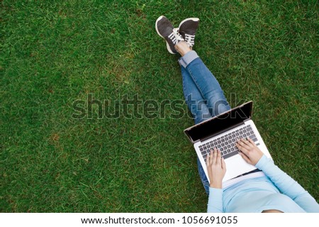 Top view of girl sitting in park on the green grass with laptop, hands on keyboard. Copy space on computer screen. Student studying outdoors.