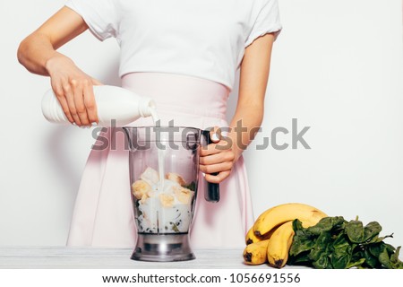 Young woman making spinach banana smoothie on white background. She pouring milk in blender and mix ingredients together. Simple elegant picture with copy space.