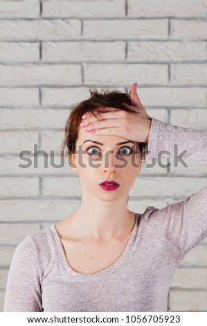 A pretty girl with surprised face, big opened eyes, her hand at her forhead, with white brick wall in the background