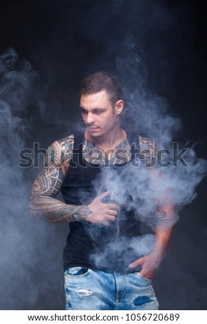 Vaper. The man with a muscular torso with tattoos smoke an electronic cigarette on the dark background