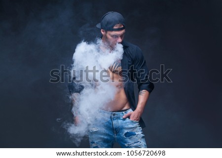 Vaper. The man dressed blue jeans, black shirt and black baseball cap with tattoos smoke an electronic cigarette on the dark background