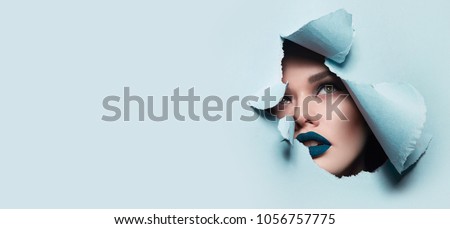 the face of a young beautiful girl with a bright make-up and puffy blue lips peers into a hole in blue paper.Fashion, beauty, make-up, cosmetics, beauty salon, style, personal care, geometry, texture. Royalty-Free Stock Photo #1056757775
