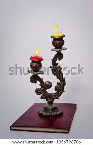 An old candlestick with a lit red and yellow candle stands on a book in front of a white background