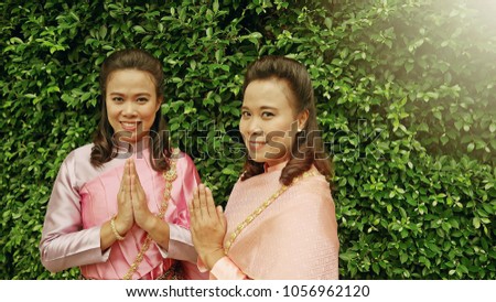 Songkran or Welcome to Thailand concept twin sisters wearing Thai traditional dress making hand sawasdee. Sawasdee is Thai greeting meaning hello. (green leaves background space for text)