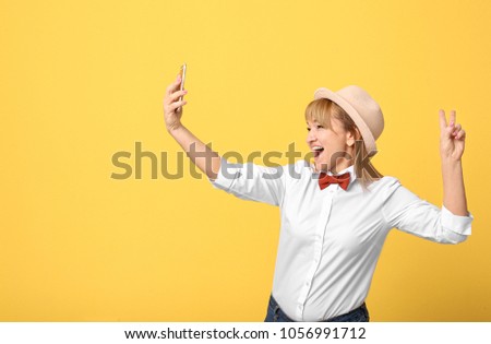 Happy senior woman in hipster outfit taking selfie on color background