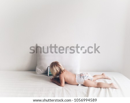 Little blonde baby girl lying at white bed watching cartoons at laptop in sunny room