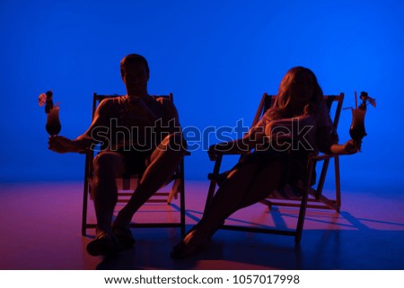 Silhouettes of man and woman holding glasses with cocktails resting in deck chairs on blue background in dark light